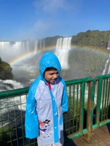 Lucas sorrindo nas Cataratas do Iguaçu com um arco-íris formado pela névoa das quedas d’água ao fundo