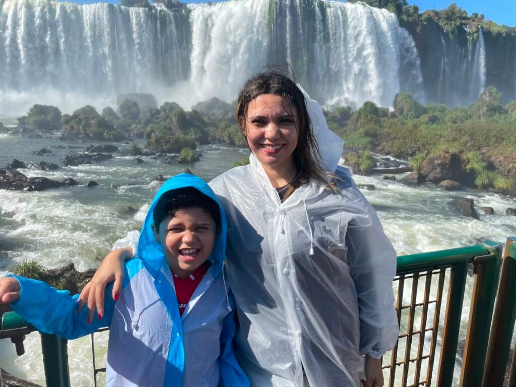 Glaziele e Lucas na passarela das Cataratas do Iguaçu, molhados pela névoa das quedas d'água durante passeio em família.