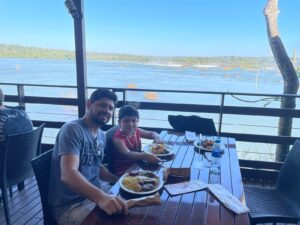 Lucas e Rafael no Restaurante Porto Canoas, nas Cataratas do Iguaçu, observando o Rio Iguaçu e a paisagem natural ao fundo. O restaurante fica às margens do rio, com uma das vistas mais bonitas do parque
