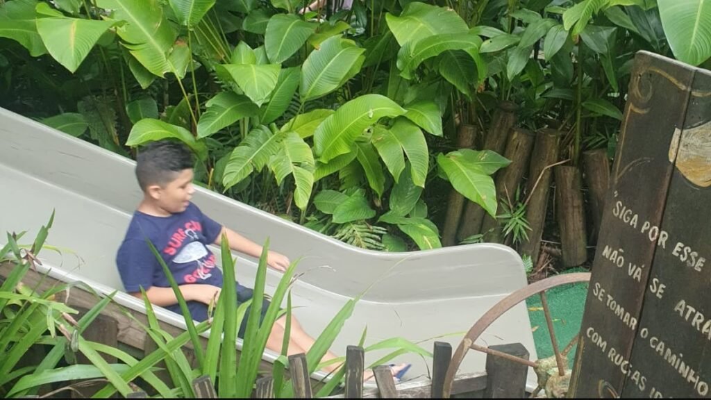 Lucas descendo pelo escorregador da Fantástica Floresta no Parque Unipraias, cercado pela vegetação.