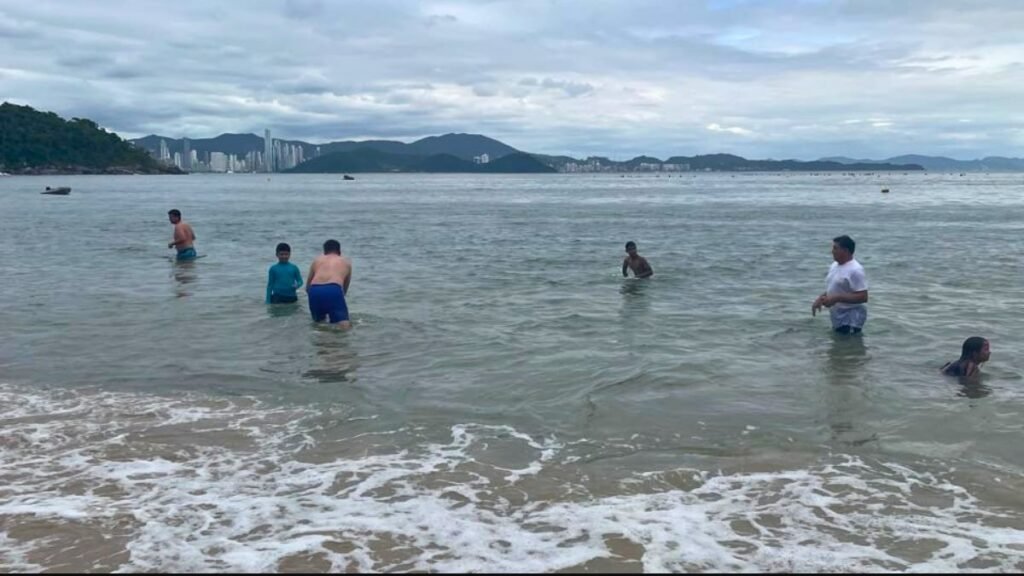 Mar de Balneário Camboriú com céu nublado, ondas suaves e prédios ao fundo.