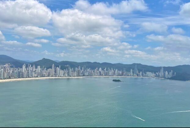 Vista panorâmica da Praia Central de Balneário Camboriú, com o mar, os edifícios altos e as montanhas ao fundo.