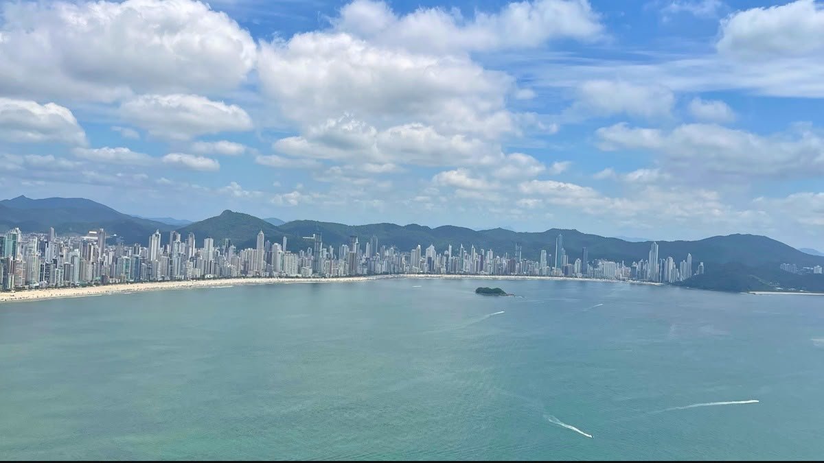 Vista panorâmica da Praia Central de Balneário Camboriú, com o mar, os edifícios altos e as montanhas ao fundo.