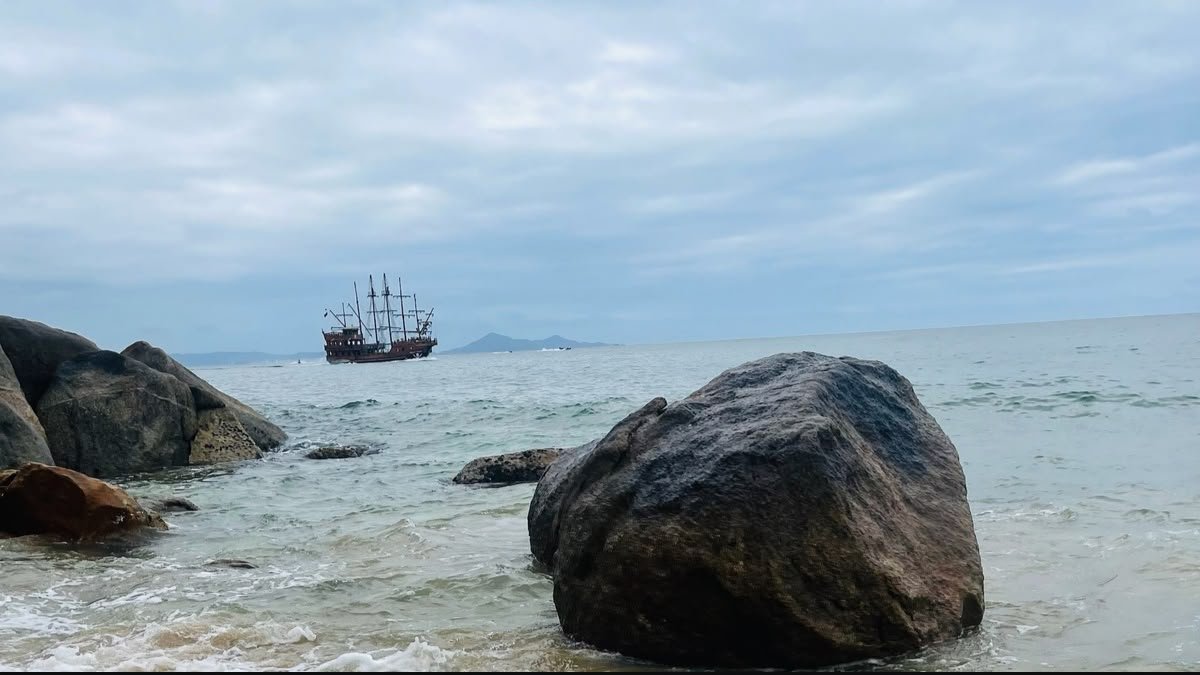 Barco turístico ao longe, mar e grandes rochas no litoral de Balneário Camboriú.