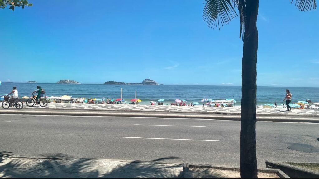 Praia de Copacabana com mar azul, banhistas, coqueiros e o calçadão de ondas em preto e branco.