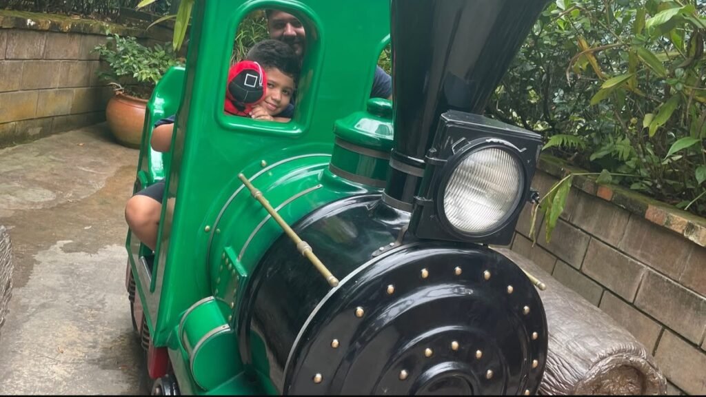 Lucas sentado em uma locomotiva verde temática na Fantástica Floresta do Parque Unipraias.